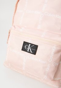 Šviesiai rožinė kuprinė, pagaminta iš nailono, su balta pakartotine ženklinimo. Yra priekinis užtrauktukas kišenėje ir juodas logotipo pleistras su baltomis raidėmis.
