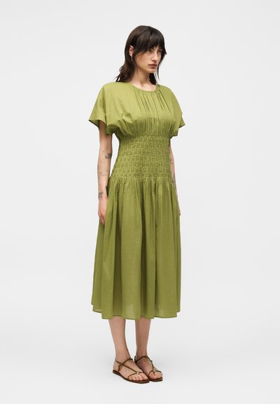 Γυναίκα που φοράει ένα ελιάς πράσινο φόρεμα μέχρι το γόνατο με σκουφάκια στη μέση και κοντά μανίκια, σε συνδυασμό με καφέ ανοιχτά σανδάλια, στέκεται σε λευκό φόντο.