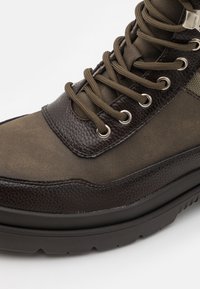Botín de tobillo de cuero marrón oscuro con materiales texturizados, diseño de cordones prominente, ojales de plata y suela de goma gruesa.
