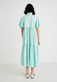 Vestido de algodón en verde menta con volante, cuello alto y mangas con volantes. Combinado con zapatos plateados, se muestra desde atrás.