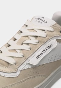 Gros plan sur une basket en daim beige et blanc avec des lacets blancs et le logo "Copenhagen Studios" sur la languette et le panneau latéral.