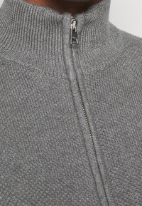 Sudadera gris con cremallera, que presenta un cuello acanalado y una tela texturizada. Detalle de cremallera metálica en la parte frontal que añade funcionalidad.