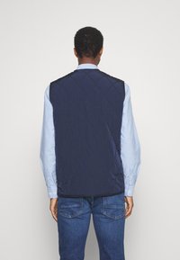 Marineblau gesteppte Weste mit glatter Textur, hohem Kragen und Reißverschluss, über einem hellblauen Hemd getragen, von hinten gesehen.