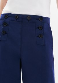 Granatowe spodnie z wysokim stanem, ozdobione dwoma rzędami czarnych guzików oraz kieszeniami po bokach. Materiał ma gładką teksturę i dopasowany krój.