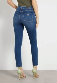 Jeans skinny din denim albastru închis, cu buzunare la spate, cusături curbe și un detaliu în formă de triunghi. Acționați cu tocuri verzi.
