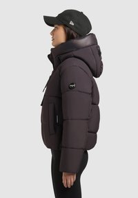 Puff jakke i mørk lilla med stor hætte, lynlåslukning og en sidelomme. Kombineret med en sort kasket og sorte leggings, vist fra siden.