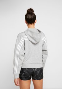 Šedá mikina s kapucí a bílými pruhy podél rukávů. Má volný střih a žebrované manžety. V kombinaci s černými vzorovanými šortkami.