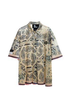 Joven de pie con camisa de manga corta beige con estampado, pantalones cortos a cuadros verde claro con ribete negro, calcetines blancos y zapatillas beige.