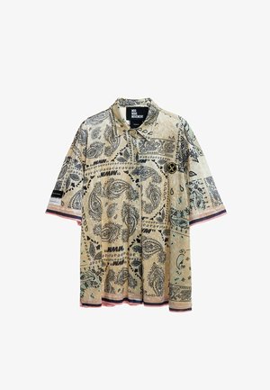 Joven de pie con camisa de manga corta beige con estampado, pantalones cortos a cuadros verde claro con ribete negro, calcetines blancos y zapatillas beige.