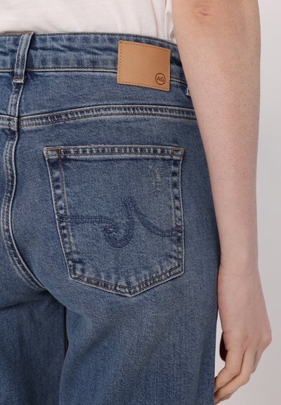 Denim jeans v srednji modri barvi z gladko teksturo. Imajo usnjeno nalepko z napisom "AG" in zadnje žepke z modrim šivom.