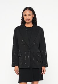 Blazer nero con borchie argento, chiusura a un bottone, risvolti a lancia e due tasche frontali. Forma strutturata con maniche lunghe.