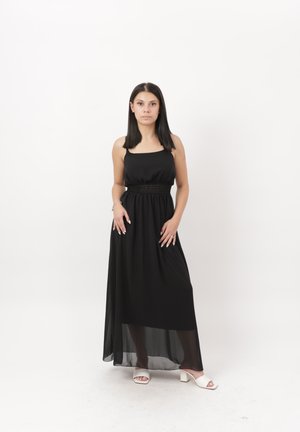 Mujer con cabello oscuro y liso lleva un vestido negro largo sin mangas y sandalias blancas de tacón ancho con punta abierta, de pie sobre un fondo blanco.