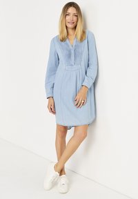 Lichtblauwe denim jurk met lange mouwen, knoopsluiting, geplooid voorpand en een licht gebogen zoom. Ggedragen met witte sneakers.