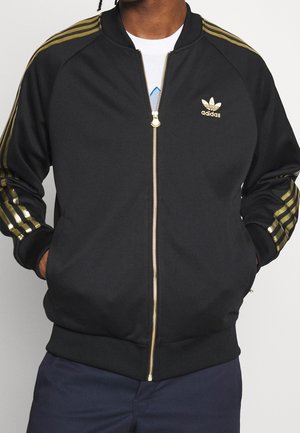 Personne portant une veste Adidas noire avec des bandes dorées sur les manches et une fermeture éclair dorée, mains dans les poches, et un pantalon bleu marine.