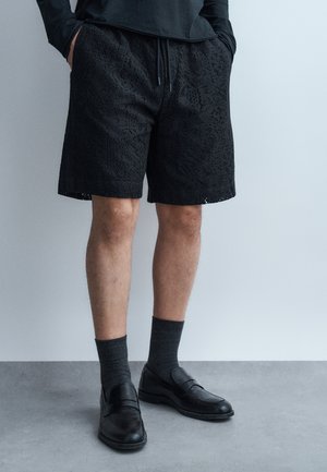 Muž v čiernych textúrovaných šortkách, tmavosivých ponožkách a čiernych kožených mokasínach stojí opretý o svetlosivú stenu a podlahu.