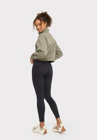 Suda verde olivo con cuello alto, combinado con leggings negros y zapatillas beige con detalles marrón claro.