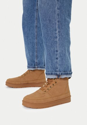 Beżowe trampki za kostkę wykonane z płótna z metalowymi oczkami, gumową podeszwą i kontrastującym przeszyciem, zestawione z niebieskimi dżinsami.