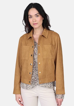 Femme aux cheveux foncés portant une veste marron clair boutonnée par-dessus un chemisier à imprimé serpent et un pantalon de couleur claire, debout devant un fond blanc.