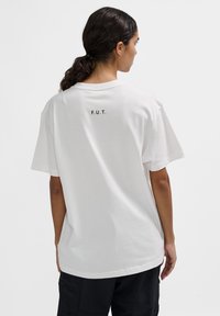 Biała bawełniana koszulka z krótkim rękawem, luźny fason, z napisem "F.U.T." nadrukowanym na czarno na górnej części pleców.