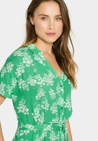 Rochie florală verde cu un design cu nasturi, mâneci scurte și un cordon. Țesătura are un aspect texturat cu modele florale albe.