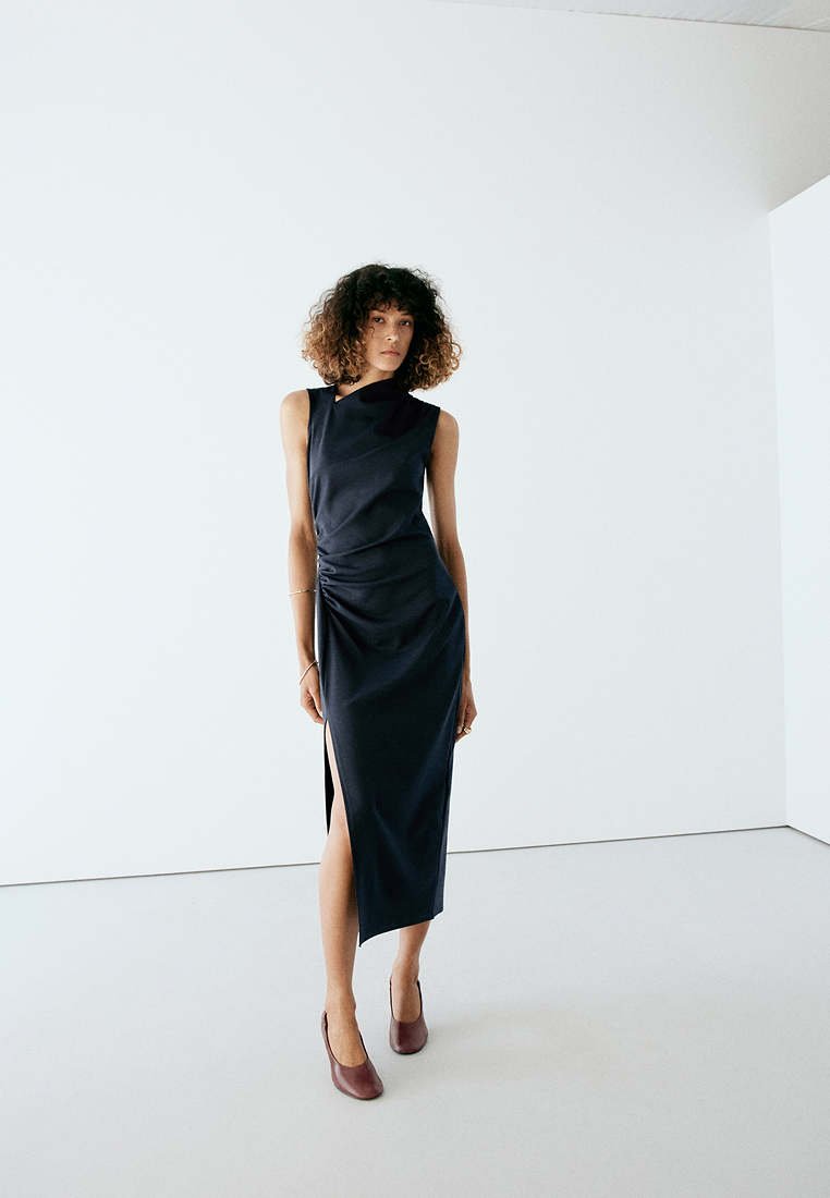 Femme aux cheveux bouclés portant une robe noire sans manches avec une fente sur le côté et des chaussures marron, debout dans une pièce blanche minimaliste.