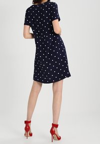 Námořnický modrý šaty s puntíky, krátké rukávy, volný střih a délka po kolena, doplněné červenými sandály na vysokém podpatku.