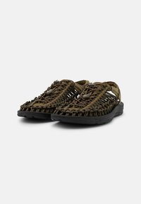 Åpne tå sko med en vevd brun tekstiloverdel, mørk gummisåle og elastiske detaljer for en sikker passform. Har utskjæringer for ventilasjon.