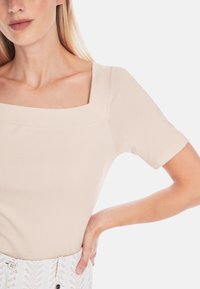 Light beige square-neck top made of smooth fabric, featuring short sleeves and a fitted design, paired with patterned white high-waisted bottoms.