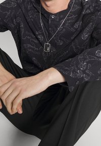 Zwart overhemd met tijgerpatroon, zilveren ketting en donkere broek. Het overhemd heeft een subtiele textuur en een getailleerd ontwerp.