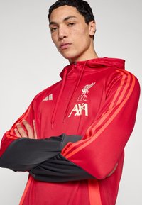 Červená sportovní bunda s kapucí a černými akcenty, s logy L.F.C. a AXA, oranžové pruhy na rukávech a zip na přední straně.