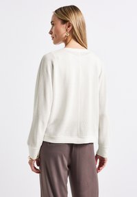 Lehká bílá mikina s volnými rameny a žebrovaným lemem, v kombinaci s hladkými, tmavě hnědými kalhotami. Jednoduchý design, volný střih.