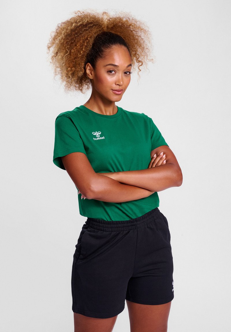 Femme aux cheveux bouclés attachés en arrière, debout, les bras croisés, portant un t-shirt vert Hummel et un short de sport noir sur un fond uni.