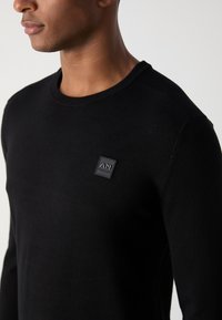 Czarny dzianinowy sweter z okrągłym dekoltem, z małą kwadratową naszywką z logo na piersi. Gładka tekstura i dopasowany krój.