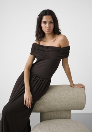 Kvinde med langt mørkt hår iført en sort off-shoulder kjole, hvilende på en moderne beige polstret bænk mod en ensfarvet grå baggrund.