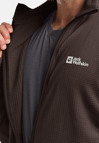 Veste zippée en marron avec une finition texturée et un t-shirt gris en dessous, arborant le logo Jack Wolfskin sur la poitrine gauche.