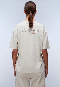 Beige bomulds T-shirt med grafik på ryggen, der viser en skiløber, korte ærmer og en afslappet pasform. Glat tekstur, enkelt design.