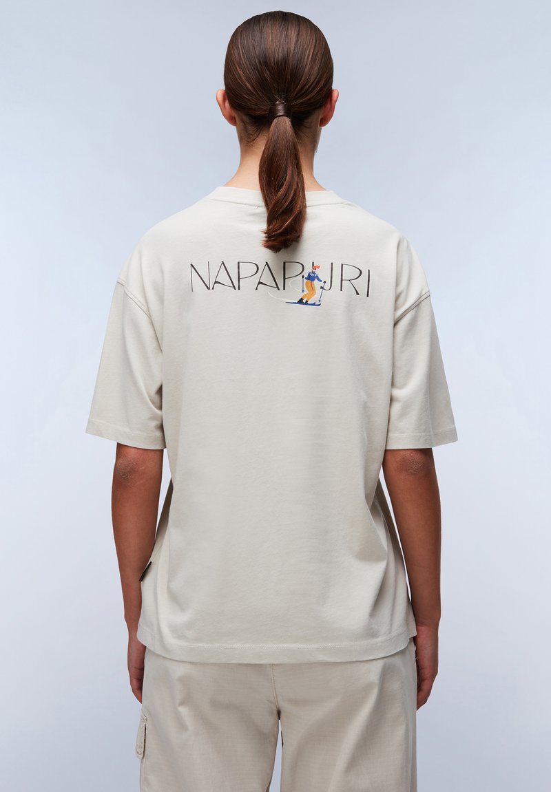 Beige bomulds T-shirt med grafik på ryggen, der viser en skiløber, korte ærmer og en afslappet pasform. Glat tekstur, enkelt design.