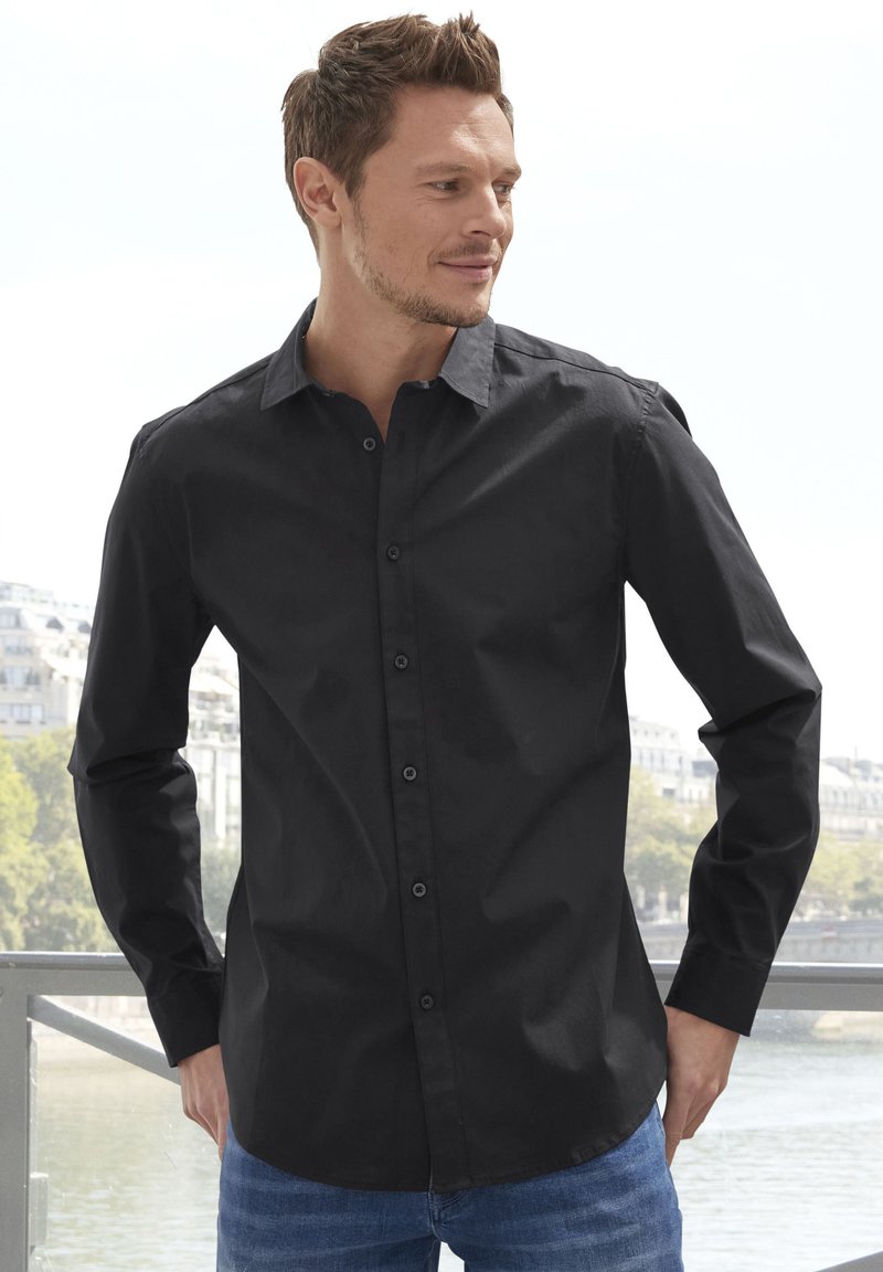 Chemise noire à manches longues, avec un col classique et une texture lisse, associée à un jean bleu. Conception simple, sans motifs ni accents.