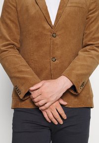 Hnědý kordový blazer s klopami, dvěma knoflíky a dekorativními manžetami. Hladká textura s vertikálními žebrovacími detaily.