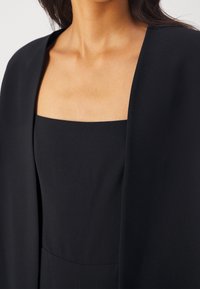 Femme portant une robe noire sans manches à encolure carrée et une veste noire assortie ouverte sur le devant, vue des épaules à la taille.