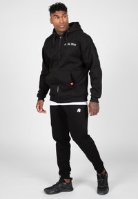 Melns uzvalka kapucis džemperis ar kangaroo kabatām, balts logo uz krūtīm; apvienots ar atbilstošām melnām sporta biksēm ar mazu baltu akcenta logo.