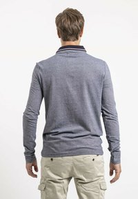 Polo shirt gris à manches longues avec un tissu texturé, présentant un col sombre avec des rayures rouges et blanches. Pantalons cargo de couleur claire visibles en dessous.