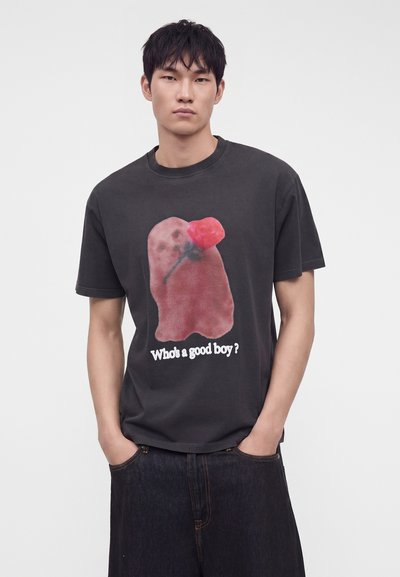 Νέος άντρας που φορά μαύρο T-shirt με θολό κόκκινο φάντασμα που κρατά μια τριανταφυλλιά και το κείμενο "Who’s a good boy?" στέκεται με τα χέρια στις τσέπες.