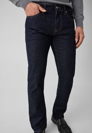 Personne portant un jean bleu foncé et des mocassins noirs, une main dans la poche, pull gris visible sur le haut du corps.