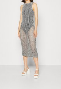 Stříbrné šaty s otevřeným pletením, kulatým výstřihem a bezrukávovým designem, dosahující délky po lýtka. Spárováno s bílými sandály na platformě.