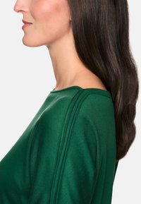 Grüner Pullover mit strukturiertem Finish, ausgestattet mit einem weiten Halsausschnitt und markanten Schulternähten, gezeigt im Profil eines Modells.