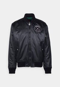 Black bomber jacket made of smooth nylon with a green lining, featuring a round white logo and two side pockets with ribbed cuffs.