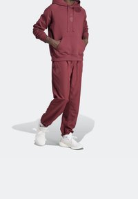 Personne portant un sweat à capuche bordeaux et un pantalon de jogging assorti avec des baskets blanches, debout les mains dans les poches du sweat à capuche devant un fond uni.