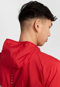 Veste légère à capuche rouge avec une texture lisse, des coutures visibles et un design minimal. Elle comporte une capuche et un détail de ventilation à l'arrière.