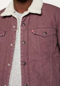 Lila denimjacka med en krämfärgad sherpa-krage, med metalltryckknappar och två bröstfickor. Grå skjorta synlig under.